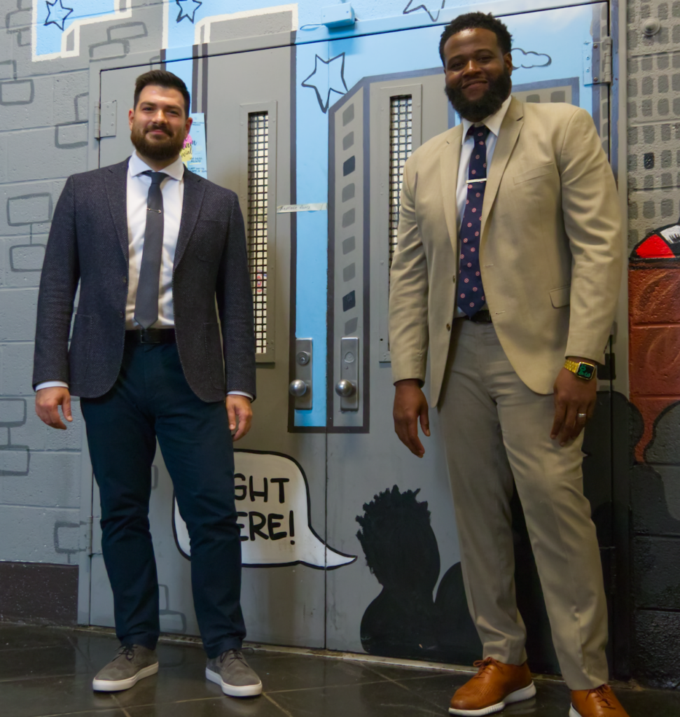 Assistant Principal Borislav Ben on the left in a dark grey suit smiles alongside Principal William Lawrence on the right in a tan suit. They are both smiling in front of the school's double doors.